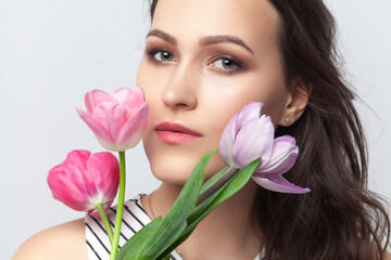 Portrait of charming romantic brunette woman looking at camera, holding spring tulips, enjoying pink flowers, wearing striped dress. Indoor studio shot isolated on gray background.