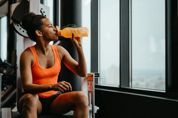 Young fit woman wearing sports clothes sitting in the fitness gym near window and drinking water...