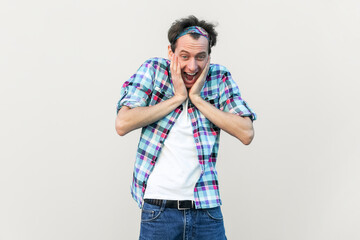 Wow, that great. Overjoyed handsome man keeps hands on cheeks, looks joyfully at camera, being amazed, wearing blue checkered shirt and headband. Indoor studio shot isolated on gray background.