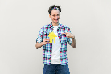 Portrait of nice attractive glad cheerful man holding in hands paper house form, sale bank loan purchase, wearing blue checkered shirt and headband. Indoor studio shot isolated on gray background.