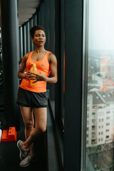 Young confident fit woman standing next to the window and looking into the distance holding water bottle resting after workout. Post workout concept.