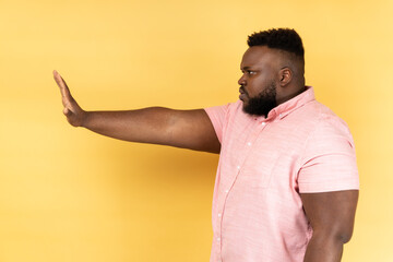 Side view portrait of dissatisfied man wearing pink shirt standing with disgust expression, frowning showing stop gesture with hand. Indoor studio shot isolated on yellow background.