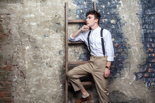 Portrait of self-confident man in white shirt and beige pants posing and lean on old wooden ladder on brick wall background, looking away, holding his chin. Indoor studio shot.