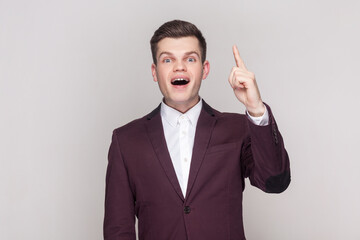 Portrait of handsome young man raises fore finger as gets idea or remembers to do something not in time, wearing violet suit and white shirt. Indoor studio shot isolated on grey background.