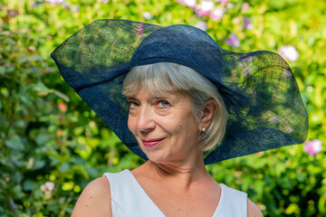Portrait of an elderly stylish woman 60-65 years old wearing a fashionable summer hat against a blurred background of nature, smiling and looking at the camera.