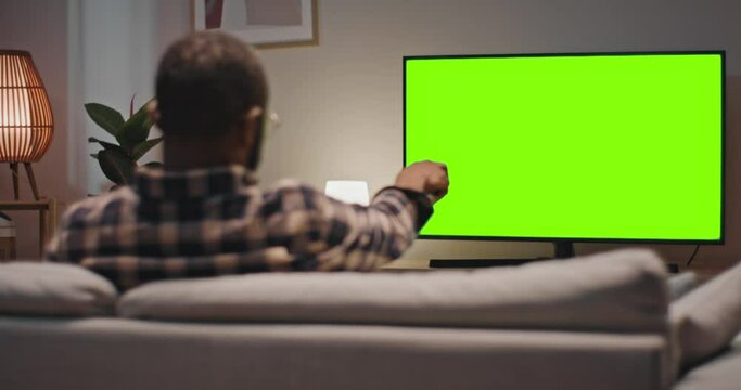 Man sitting at sofa and enjoying favourite TV show. Back view of male holding remote control and changing channel. Man relaxing sitting at couch at home. Male looking at green chroma key screen of TV.
