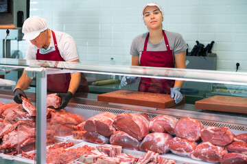 In organic natural meat store, friendly farmer female seller stands near counter refrigerator display case. In butcher shop, young female vendor in uniform apron, gloves, cap holds cutting board