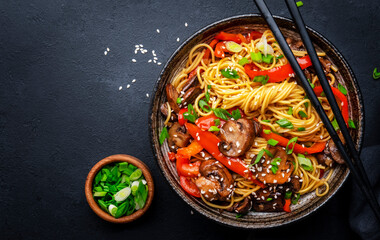 Stir fry noodles with vegetables: red paprika, champignons, green onion and sesame seeds in ceramic bowl. Black table background, top view