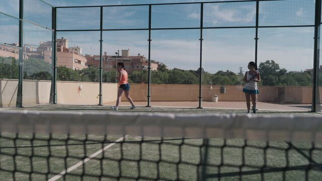 Two padel tennis women players hit back the ball with a drive shot after the ball hits the backwall.