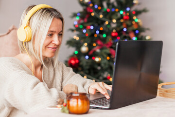 Adult woman in cozy white knitted sweater using laptop  in living room in christmas