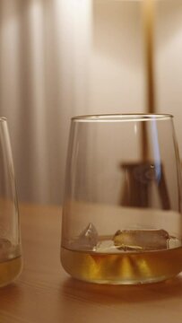 Vertical video. The man and woman place their glasses of whiskey next to each other on a wooden table. The scene is illuminated by dim and warm lighting in a white room.