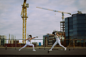 Two fencers woman practice on a rooftop with the city in the background.