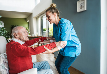 Smiling nurse helping senior man to exercise and holding hands at home