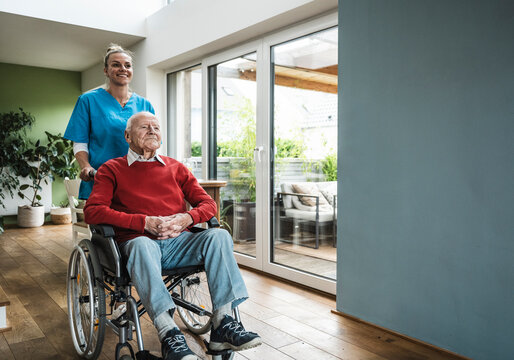 Happy nurse taking care of senior man sitting in wheelchair at home