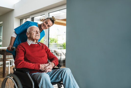 Happy nurse taking care of senior man sitting in wheelchair by wall at home