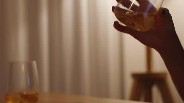 Two glasses of whiskey on a wooden table, but the young woman drinks alone in dim lamplight.