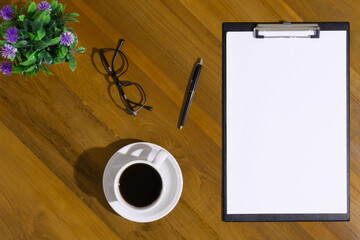 Clipboard mockup with a pen, a cup of coffee, a pair of glasses and an artificial plant on top of wooden desk background
