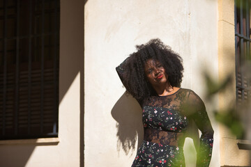 Young woman, beautiful and black with afro hair, with floral dress leaning on a wall, relaxed and quiet, seen through a green plant. Concept beauty, relaxation, tranquility, plants.