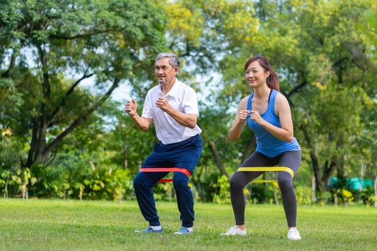 Senior asian man and his daughter are using sport rubber band to build up his leg muscle strength  in the public park for elder longevity exercise and outdoor workout
