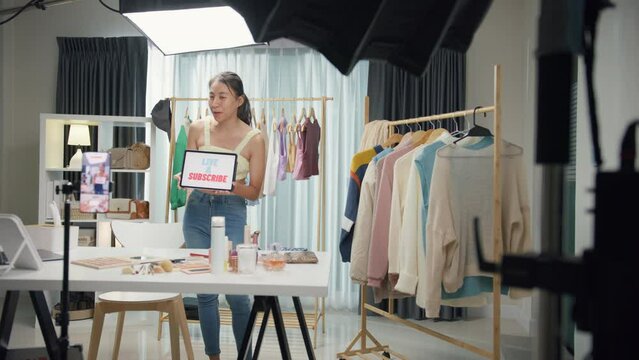 Young Asia lady beauty blogger wear crop top looking at phone camera live stream use digital tablet talk follower introduce her chanel ask audience like subscribe share her chanel at house studio.