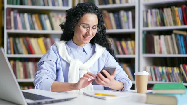 Smiling female student using smartphone sitting in campus library space. Happy girl looking for information on the internet or chatting with a friend, writing a text message, browsing social networks