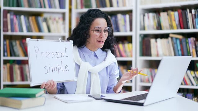 Smiling female teacher in glasses giving video call English lesson using laptop sitting in campus library space. A positive woman tutor or coach has online training, conducts a course, talks remotely