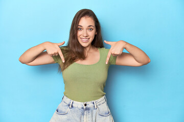 Fototapeta premium Young Caucasian woman in a green top on a blue backdrop points down with fingers, positive feeling.