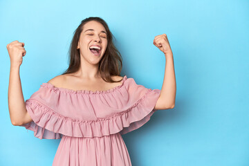 Fototapeta premium Young woman wearing a pink dress on a blue studio backdrop raising fist after a victory, winner concept.