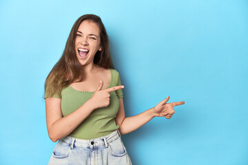 Young Caucasian woman in a green top on a blue backdrop pointing with forefingers to a copy space, expressing excitement and desire.