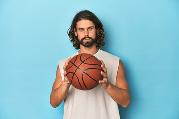 Long-haired, bearded man holding a basketball in a blue studio.