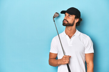 Bearded, long-haired golfer holding a golf club in a blue studio.