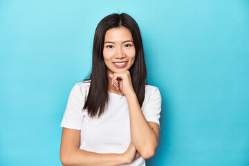 Young Asian woman in white t-shirt, studio shot, smiling happy and confident, touching chin with hand.