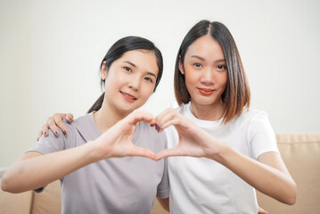 Affection LGBT concept. Happy diverse beautiful asian female lesbian couple making heart shape with hands together on sofa at home. Spending quality time together.