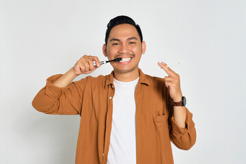 Happy young Asian man in casual shirt brushing his teeth with toothbrush while holding cigarette isolated on white background