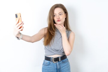 Young beautiful girl wearing grey T-shirt blows air kiss at camera of smartphone and takes selfie, sends mwah via online call.