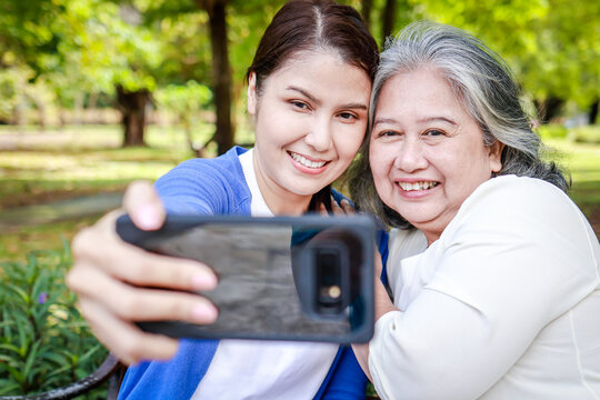portrait of an elderly mother with her daughter Sit and relax on a bench in the park. They were smiling happily holding their smartphones taking pictures together. Family concept. health care, life in