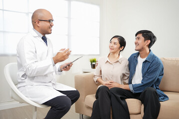 Male doctor giving test result to asian couple patient with tablet. They feeling happy and holding hand together when received good news from the doctor in medical office, hospital, clinic or home.