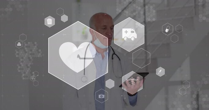 Animation of multiple icons over senior caucasian male doctor looking at digital tablet in hospital