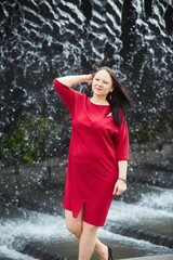 A spectacular older woman in a bright red dress on the background of a waterfall in the park.