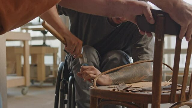 Medium shot of hardworking young Caucasian wheelchaired carpenter using hand tools to disassemble seat of old wooden chair for its further restoration, working in carpentry workshop with workmateMediu