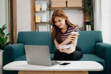 Female students note from the books at the Asian girl library sitting at sofa using laptop computer and tablet to search an online informations.