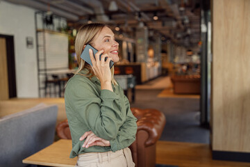 Smiling businesswoman is talking phone with client while standing on modern coworking background