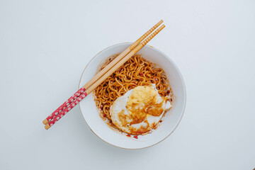 Flat lay or top view shot of a portion of stir-fried Indonesian instant noodles with egg on a white bowl with a pair of wooden chopsticks on white table