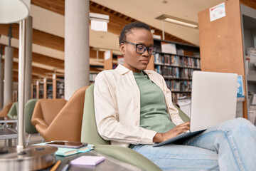 Busy short-haired African girl student using laptop technology sitting in university campus library. Serious young Black woman elearning looking at computer advertising hybrid work and elearning.