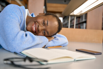 Fototapeta na wymiar Happy cute African teenage girl, smiling short-haired cute Black ethnic college student lying on desk looking at camera in modern university campus library. Copy space. Close up portrait.