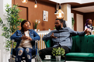 Young African American man and his disabled wife sitting in hotel lobby and talking, unhappy tired...