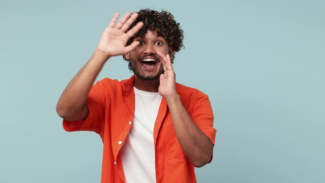 Young Indian man wear red shirt casual clothes look around for friend find waving meet greet with hand as notices someone say hello hi isolated on pastel plain blue cyan background. Lifestyle concept