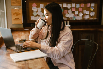 Beautiful asian woman working on laptop and drinking coffee while sitting in cafe