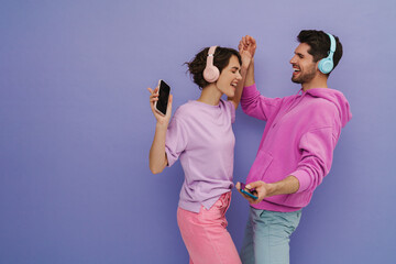 Young couple dancing while listening music with headphones and mobile phones isolated over purple...