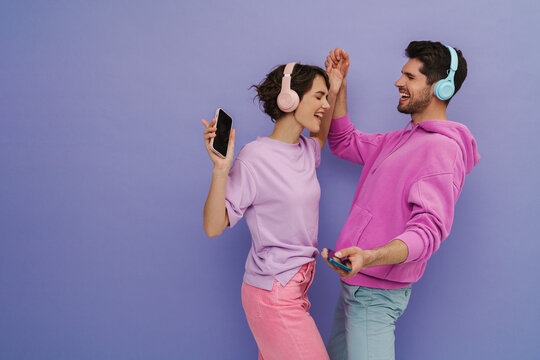 Young Couple Dancing While Listening Music With Headphones And Mobile Phones Isolated Over Purple Background